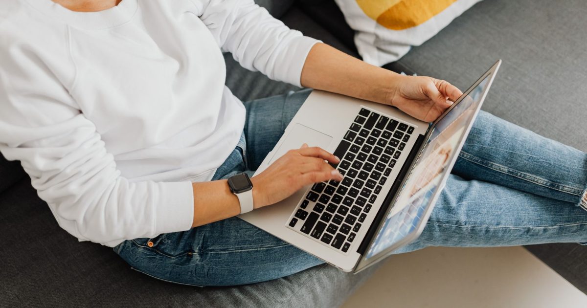 Person in casual attire using laptop on cozy grey sofa, creating a home workspace vibe.