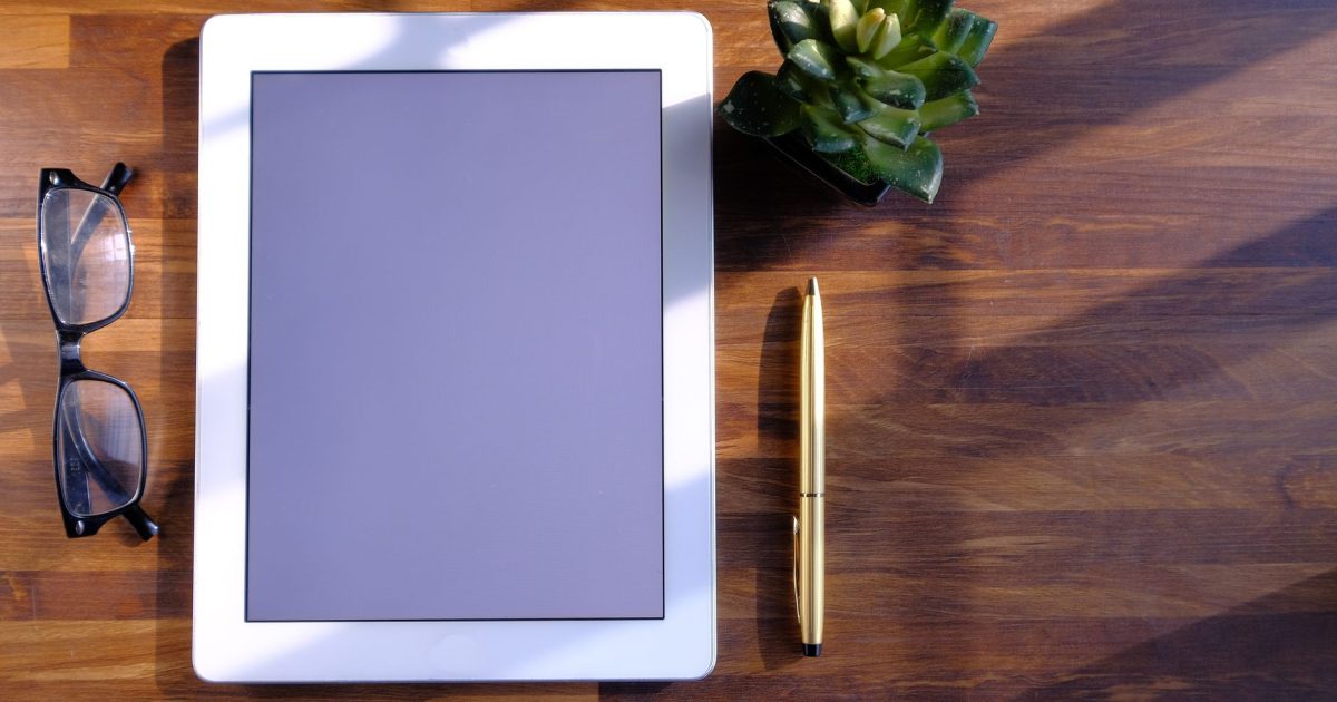 Top view of tablet, eyeglasses, pen, and succulent on wooden surface.