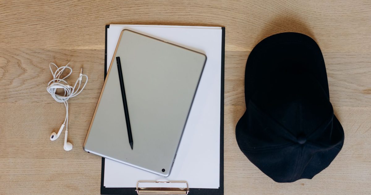 Minimalist top-down view of a digital tablet, earphones, pen, and cap on a wooden surface.