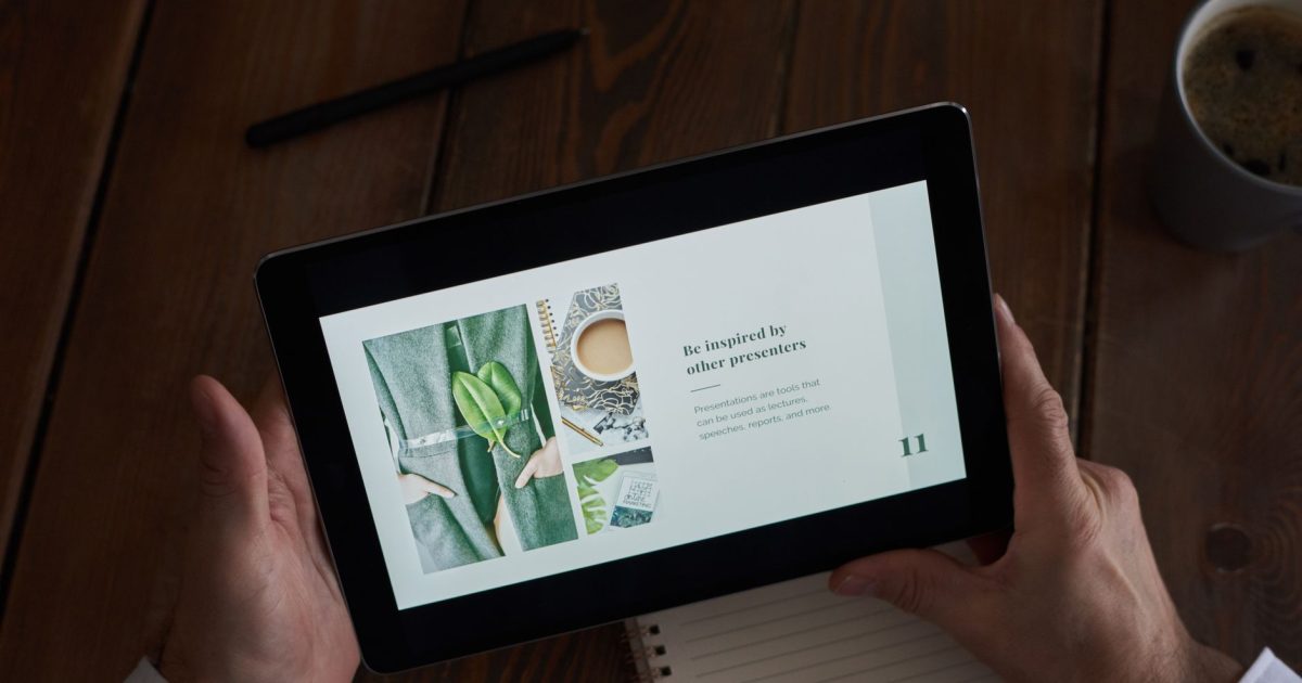 Close-up of hands holding a tablet showing a presentation on a wooden desk.