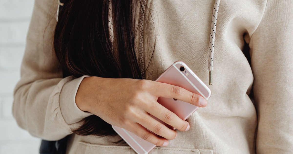 Young woman in a hoodie holding a pink smartphone indoors. Casual and modern lifestyle.