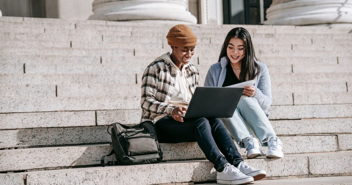 Full length of happy young multiethnic women in casual outfit with backpack sitting on steps near building while studying on computer in daylight
