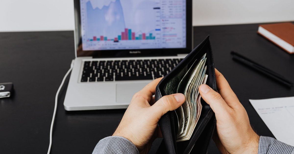 Man holding a leather wallet with bills in front of a laptop displaying financial graphs.