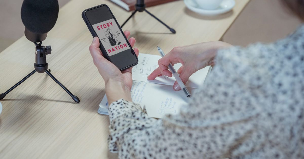 Back view faceless female in stylish blouse browsing mobile phone while sitting at desk with microphones and diary during interview