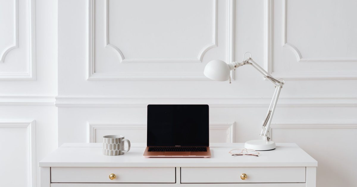 Elegant minimalist workspace with laptop and desk lamp on a white desk in a bright room.